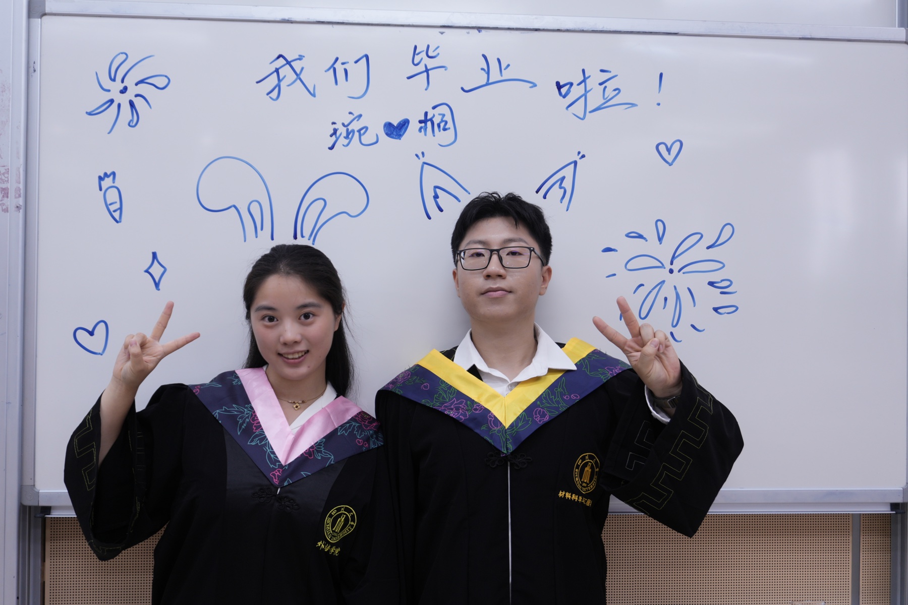 张桐宇和许琬苓穿学士服站在写有我们毕业啦的白板前